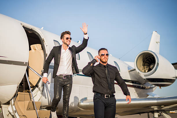 Male celebrity exiting the private jet airplane in the escort of his bodyguard dressed in black suit. Man is waving to someone who awaits.